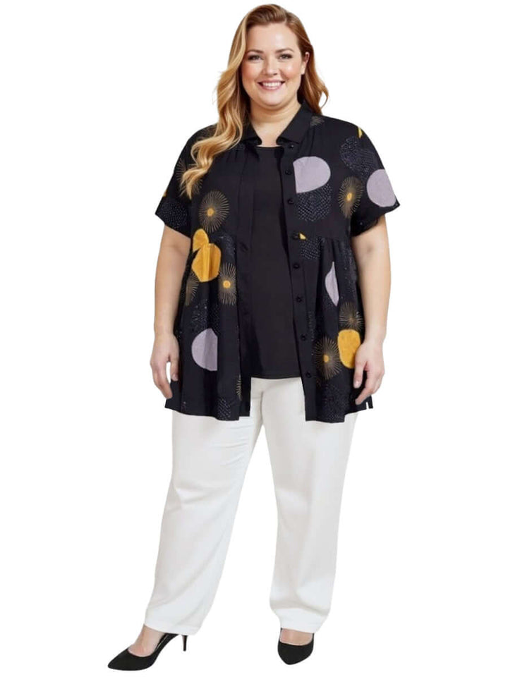Coreen Tunic - Black Print on a plus-size model, featuring a collared neckline and colorful prints, styled with white pants.